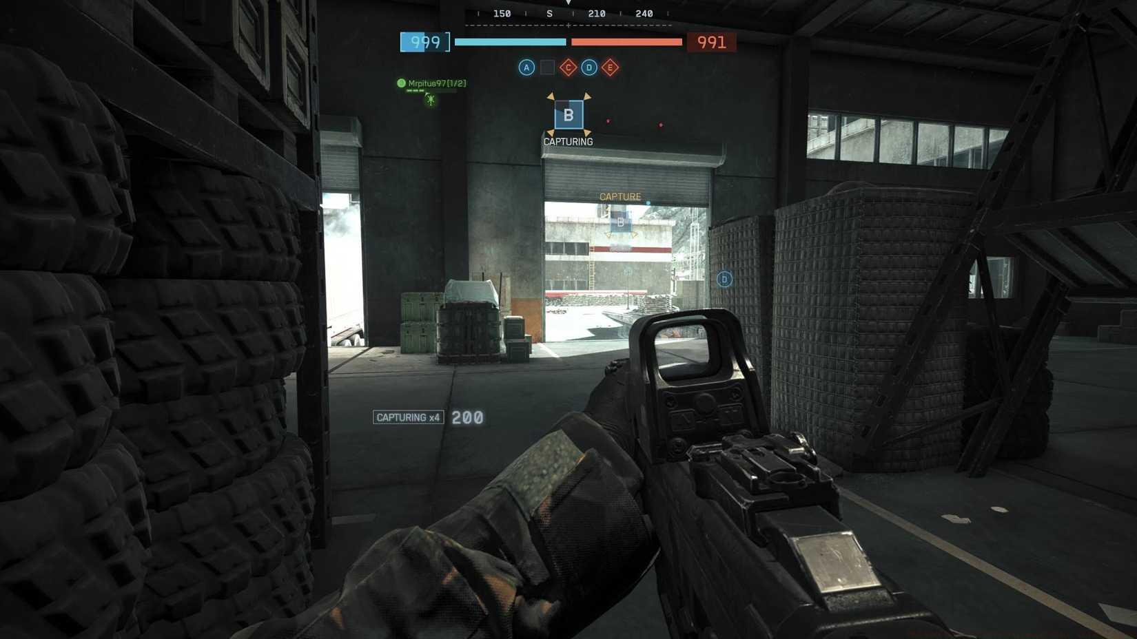 The player capturing B in Conquest on Operation Firestorm, equipped with an assault rifle hiding in a corner of a hangar.